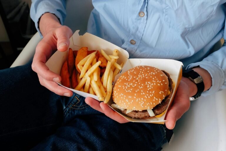 Unrecognizable male with wristwatch in casual clothes sitting on chair and holding box of burger and french fries in hands