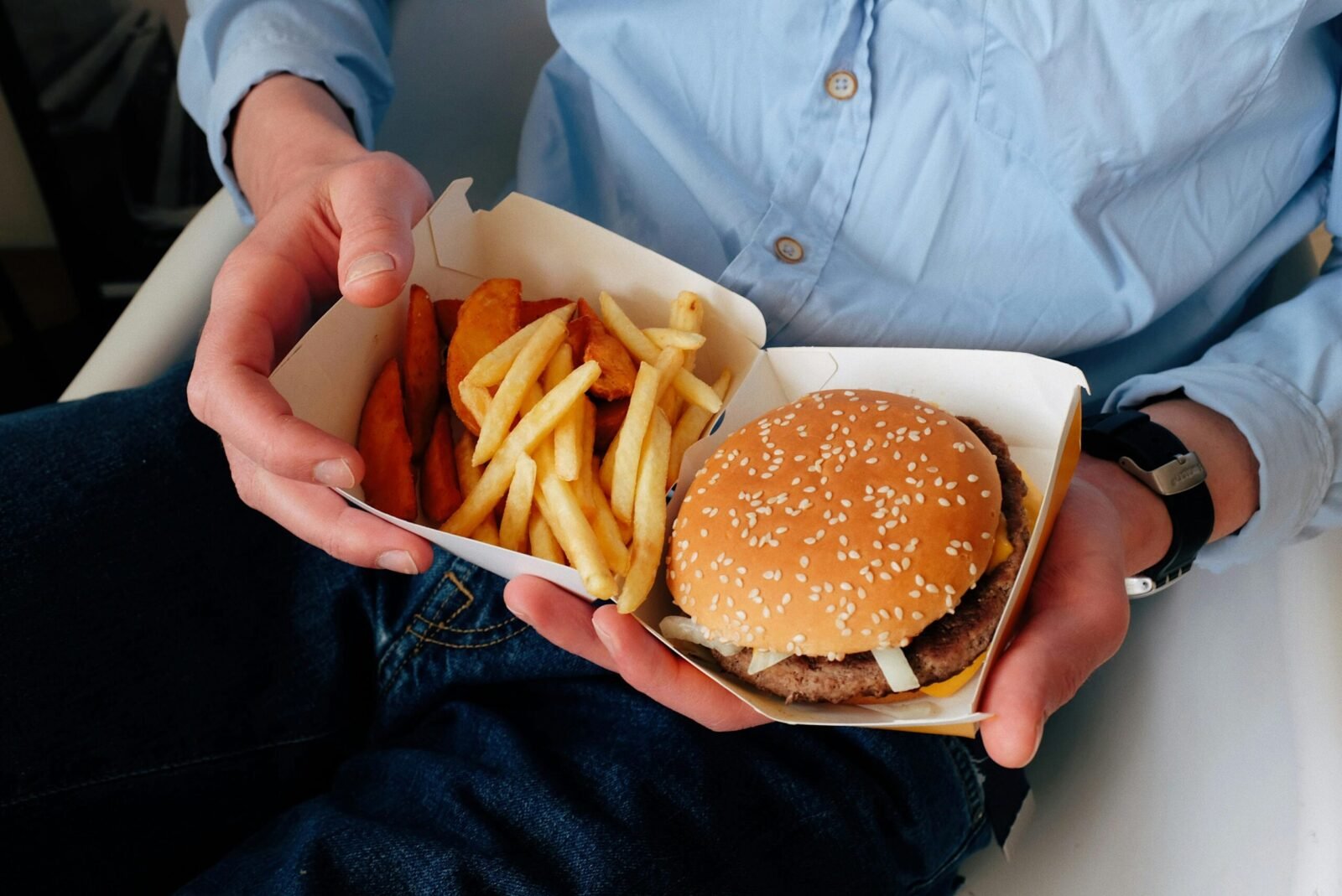 Unrecognizable male with wristwatch in casual clothes sitting on chair and holding box of burger and french fries in hands